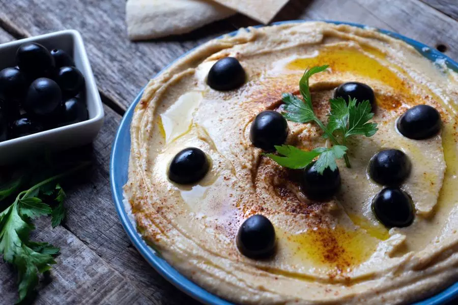 Plate of swirled hummus with olive oil, paprika, and black olives, with pita bread and a bowl of olives nearby.