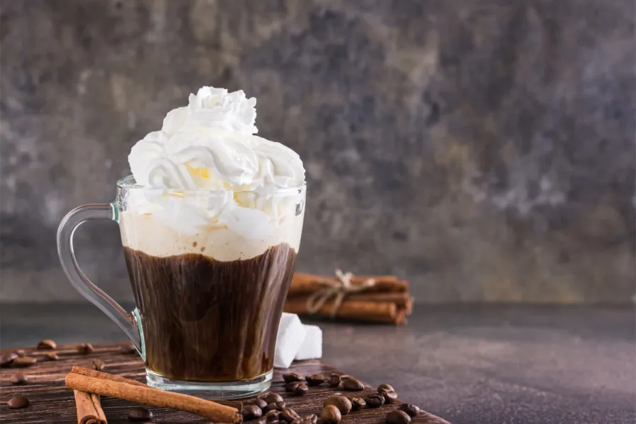 Glass mug of Einspänner coffee topped with whipped cream, surrounded by coffee beans and cinnamon sticks on a rustic table.