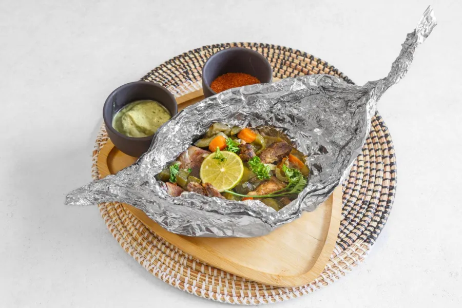 Foil-wrapped fish with vegetables and lime served on a wooden tray with dipping sauces, Congolese style