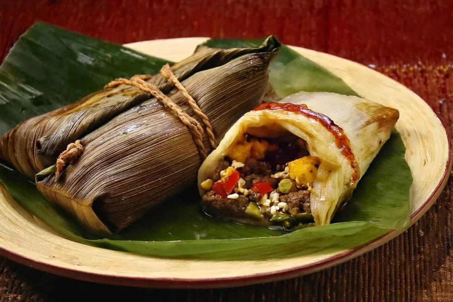 Costa Rican tamales on a banana leaf, with one tamal opened to show meat, rice, and vegetable filling.