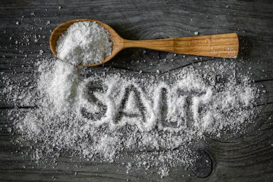 Wooden spoon filled with coarse salt on a dark table, with scattered salt spelling the word SALT.