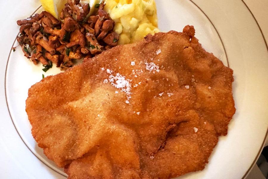 Golden breaded schnitzel with coarse salt, served with potato salad, sautéed chanterelle mushrooms, and a lemon wedge on a white plate.