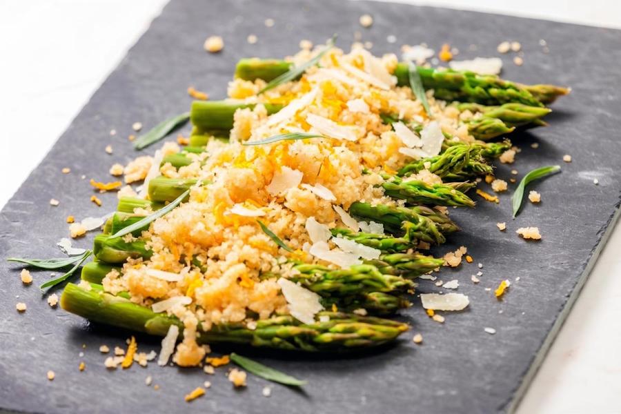 Roasted green asparagus topped with citrusy golden breadcrumbs, shaved Parmesan, orange zest, and fresh herbs on a dark slate serving board.