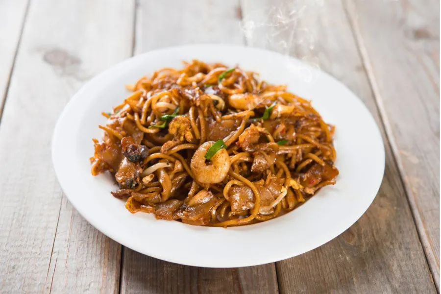 A plate of char kway teow — stir-fried flat rice noodles with shrimp, egg, bean sprouts and scallions, glistening with dark soy sauce and lightly charred, served on a white plate on a wooden table, with steam rising.