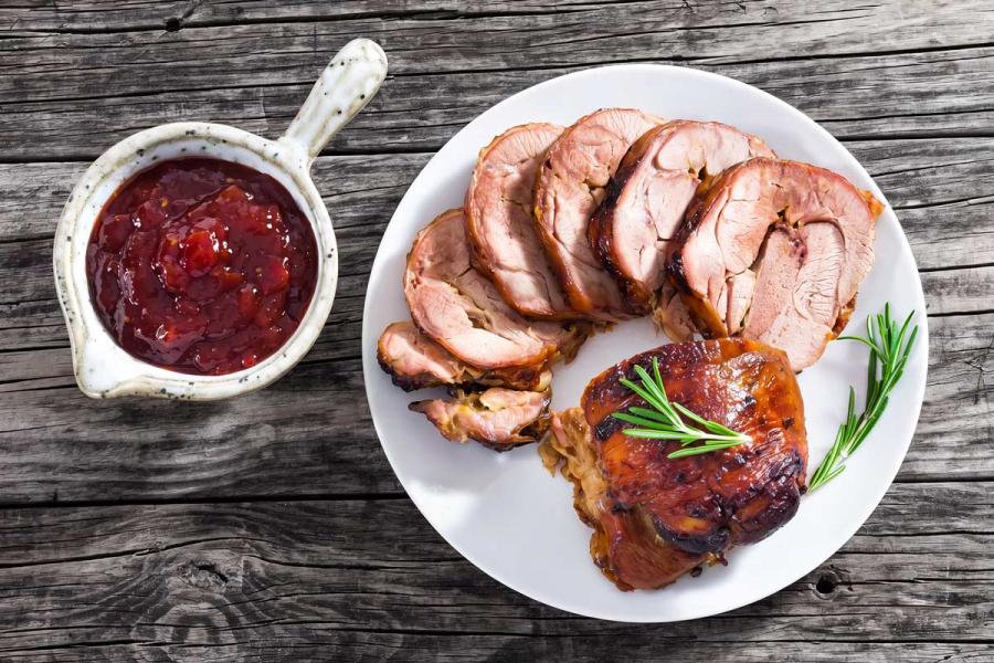 Sliced turkey roulade fanned out on a white plate with fresh rosemary, served alongside a small pitcher of chunky red chili-tomato jam on a rustic wooden table.