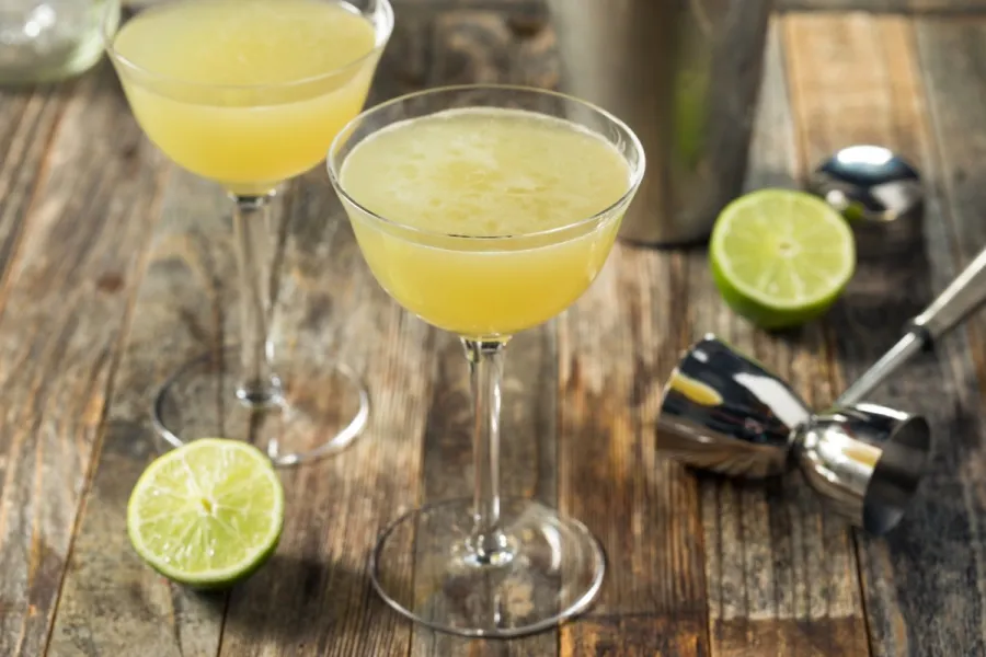 Two cloudy yellow citrus cocktails in coupe glasses on a rustic wooden table, with a halved lime, a cocktail shaker and a jigger alongside.