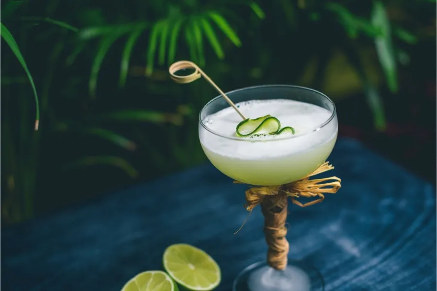 A pale green, frothy cocktail in a raffia-wrapped coupe glass, garnished with a curl of cucumber on a pick, on a blue wooden surface with lime halves and tropical foliage behind.