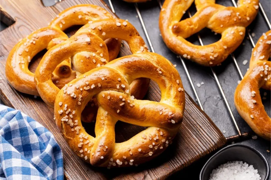 Freshly baked Bavarian pretzels scattered with coarse salt on a dark wooden board, with more cooling on a wire rack and a bowl of salt alongside