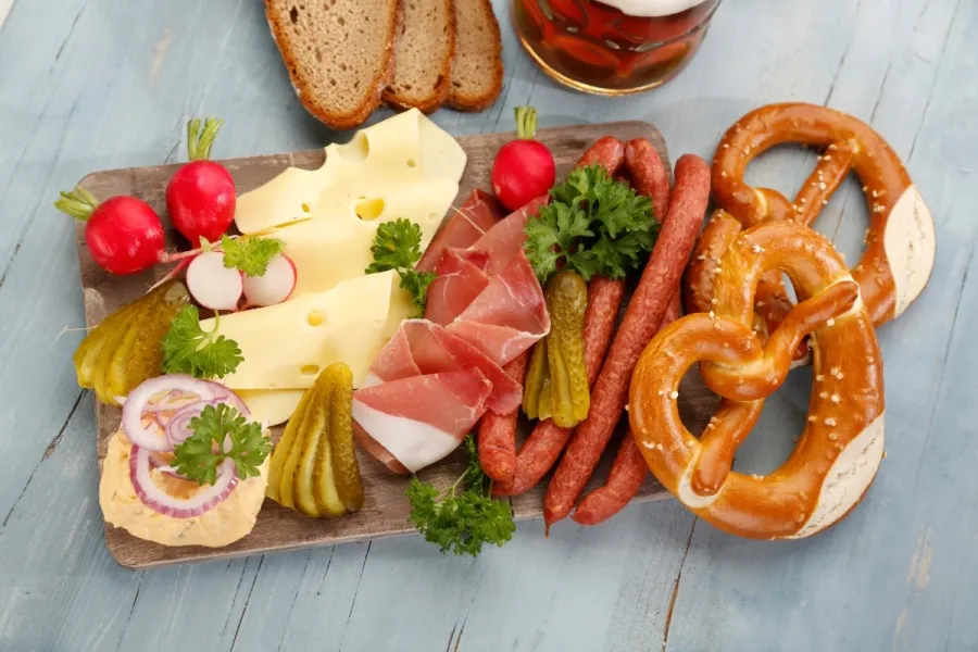 Wooden board of Bavarian cold cuts — Emmental cheese, cured ham, thin smoked sausages, gherkins, radishes and Obatzda — with two pretzels, rye bread and a beer alongside