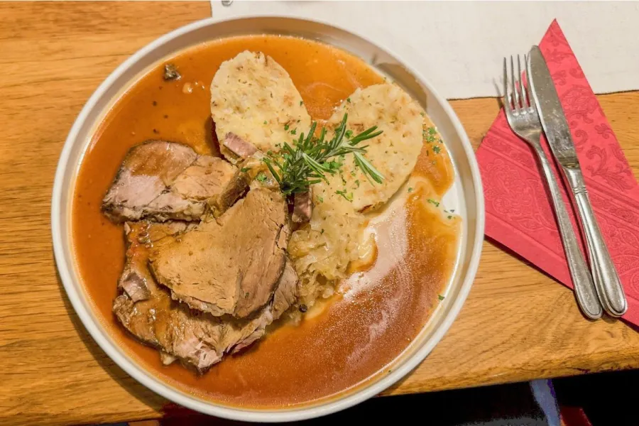 Bowl of sliced Bavarian roast pork in dark gravy with two bread dumplings, sauerkraut and a rosemary sprig, on a wooden table beside a red napkin and cutlery