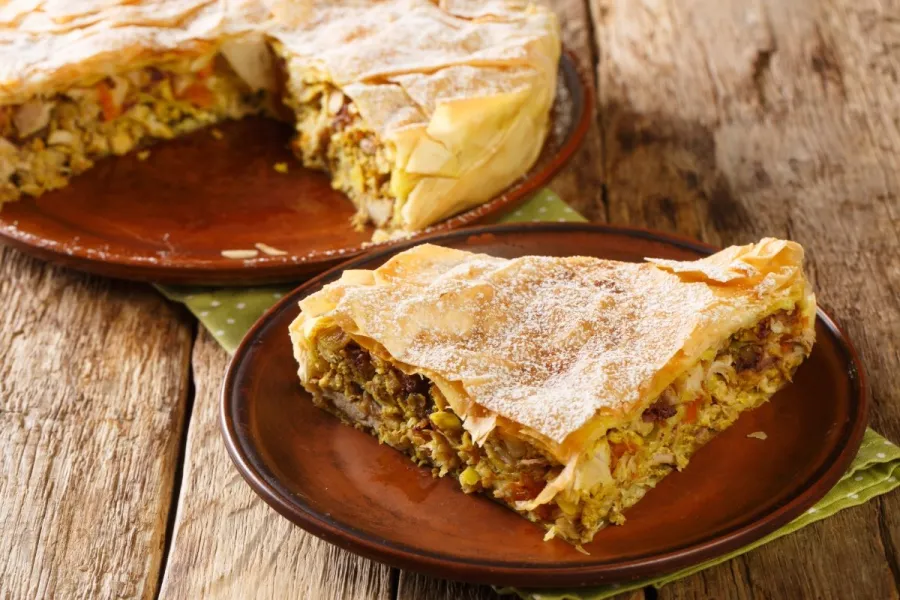 A wedge of Moroccan chicken pastilla on a rustic plate, its crisp golden phyllo crust dusted with powdered sugar, cut to reveal a spiced chicken, egg, and almond filling, with the rest of the round pie on a wooden table behind.
