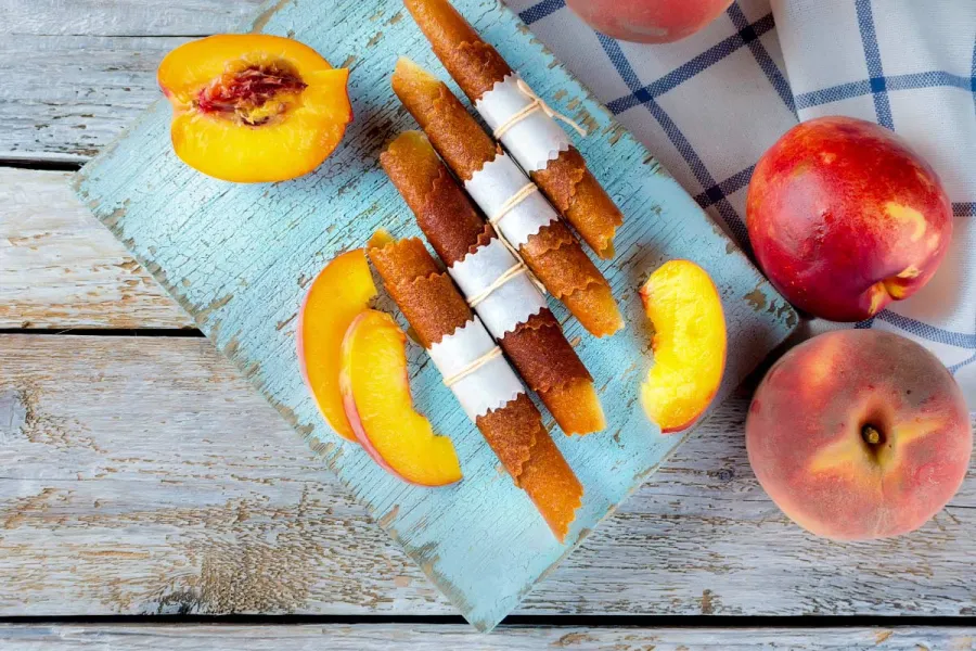 Rolled strips of peach fruit leather tied with paper and string on a pale-blue board, with fresh whole peaches, a halved peach and slices