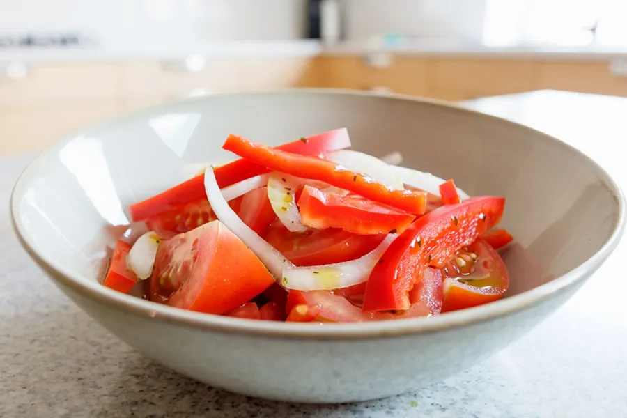 A bowl of Argentine tomato and onion salad with red bell pepper strips, dressed with olive oil and oregano