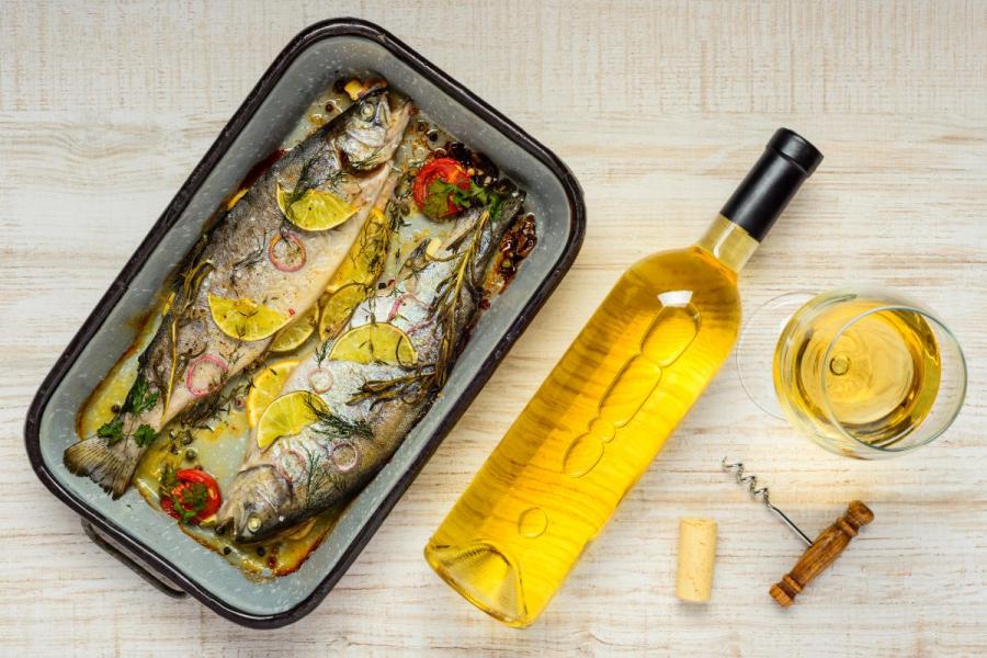 Two cooked trout in a baking dish garnished with lemon slices and herbs, accompanied by a bottle of white wine, a filled wine glass, a corkscrew, and a cork.