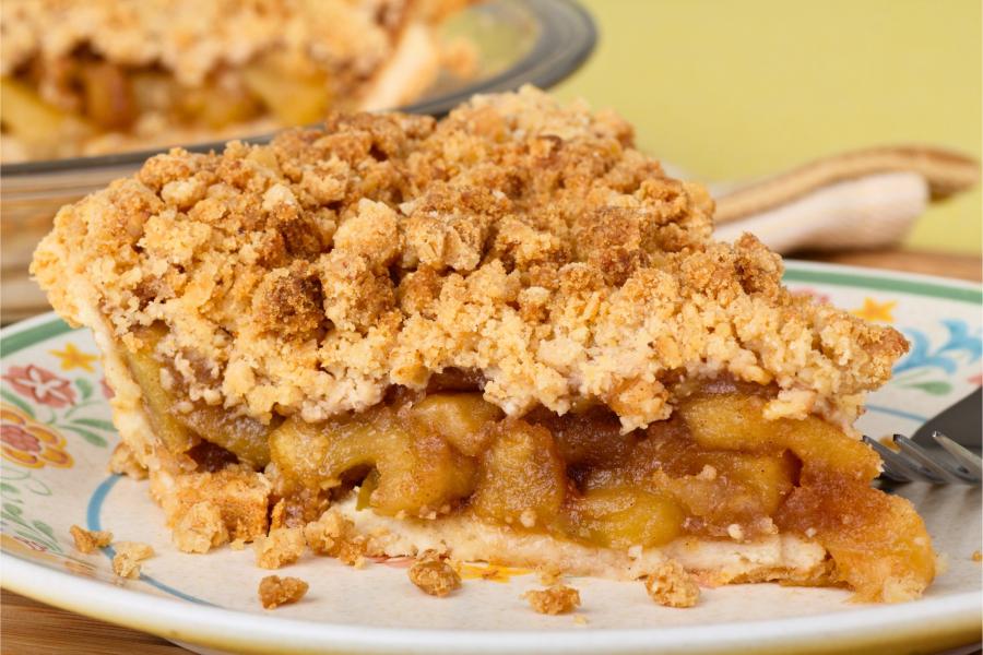 Close-up of a slice of apple crumb pie on a decorative plate, featuring a golden, crumbly topping and chunks of apple filling. The rest of the pie is visible in the background.