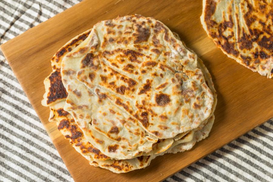 Stack of lefse, traditional Norwegian flatbread, on a wooden cutting board. The flatbreads are thin, round, and have a browned, spotted appearance. The wooden board is placed on a striped cloth.